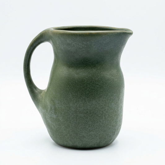 A dark green speckled stoneware pitcher with a handle and spout, sitting on a white marble countertop against a white subway tile backsplash.