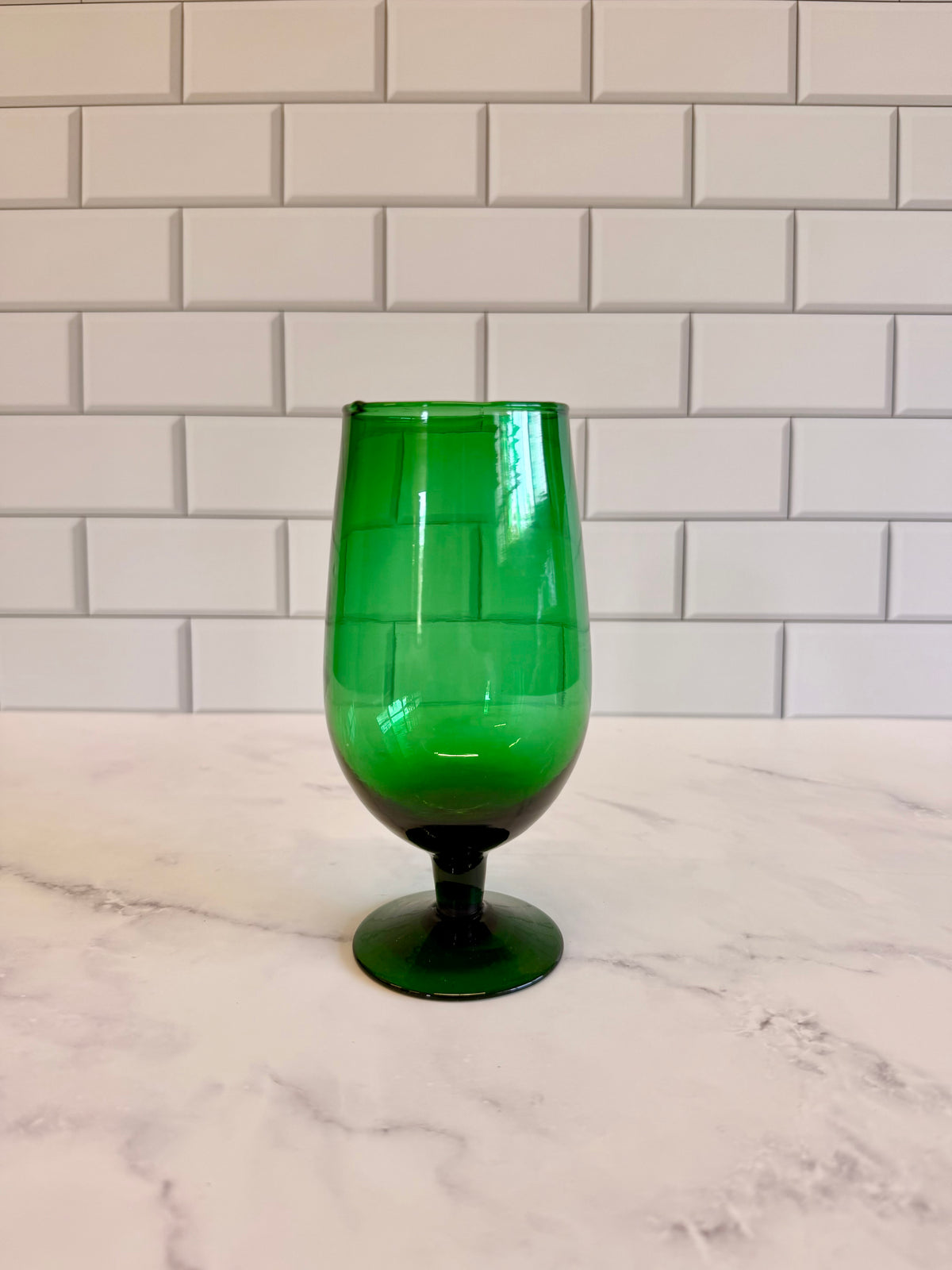A green translucent glass goblet with a wide bowl and short stem, standing on a white marble surface against a white subway tile backsplash.